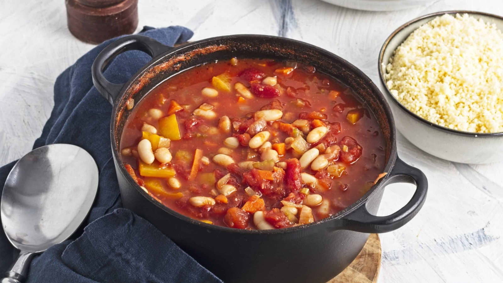 A black pot filled with a tomato-based stew containing white beans, bell peppers, and other vegetables. The pot rests on a dark cloth with a silver spoon nearby. A bowl of couscous sits beside the pot on a white surface.