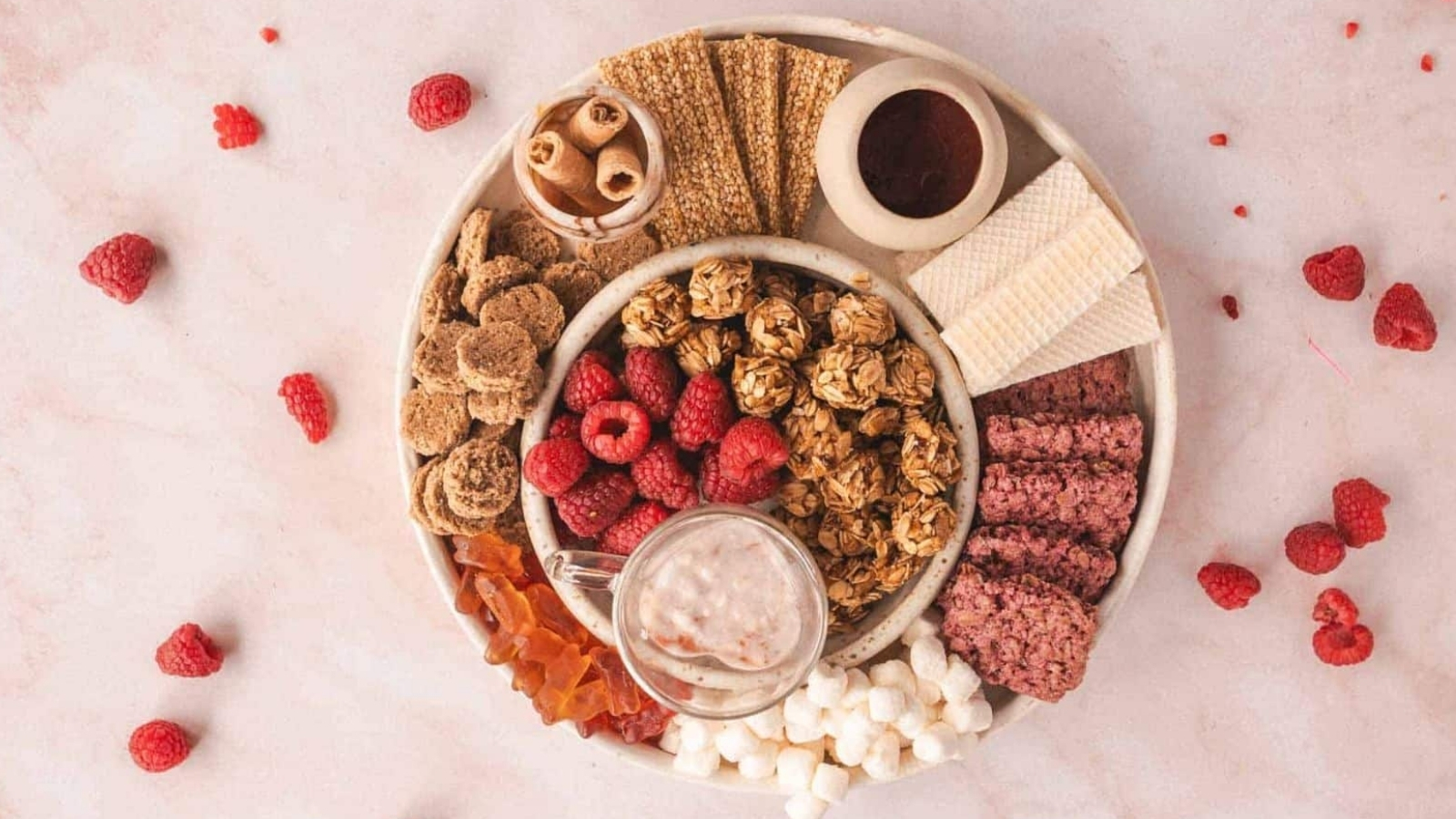 A round platter on a light surface features a variety of snacks: raspberries, granola balls, wafer rolls, crackers, a small dish of sauce, red sticks, marshmallows, and a glass cup with a light brown liquid. Scattered raspberries are around the platter.
