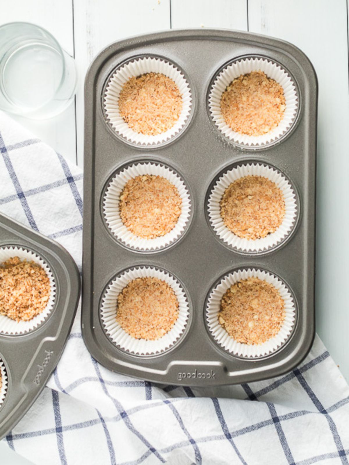 Vanilla wafer cookie crumb crust pressed into cupcake liners in a muffin pan for mini cheesecakes.