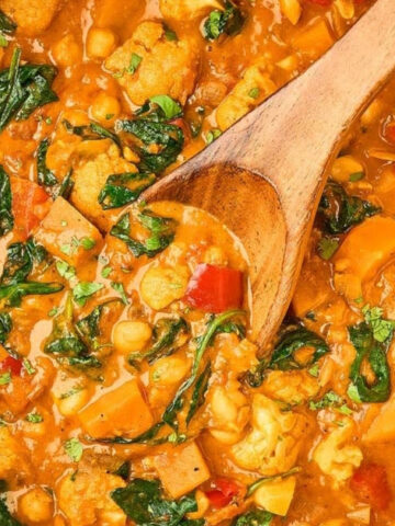 A pot filled with a tomato-based vegetable stew containing chickpeas, cauliflower, spinach, and red pepper pieces, with a wooden spoon resting inside, and bowls of rice and chopped herbs nearby.