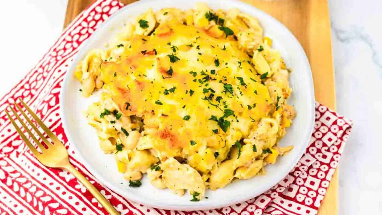 A plate of cheesy pasta casserole garnished with chopped parsley, on a red-patterned napkin with a gold fork.