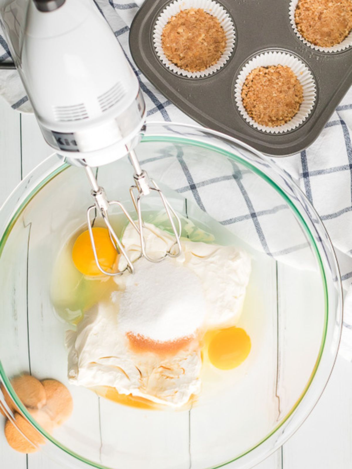 Mixing cheesecake filling with a hand mixer in a glass bowl, with graham cracker crusts in a muffin pan.