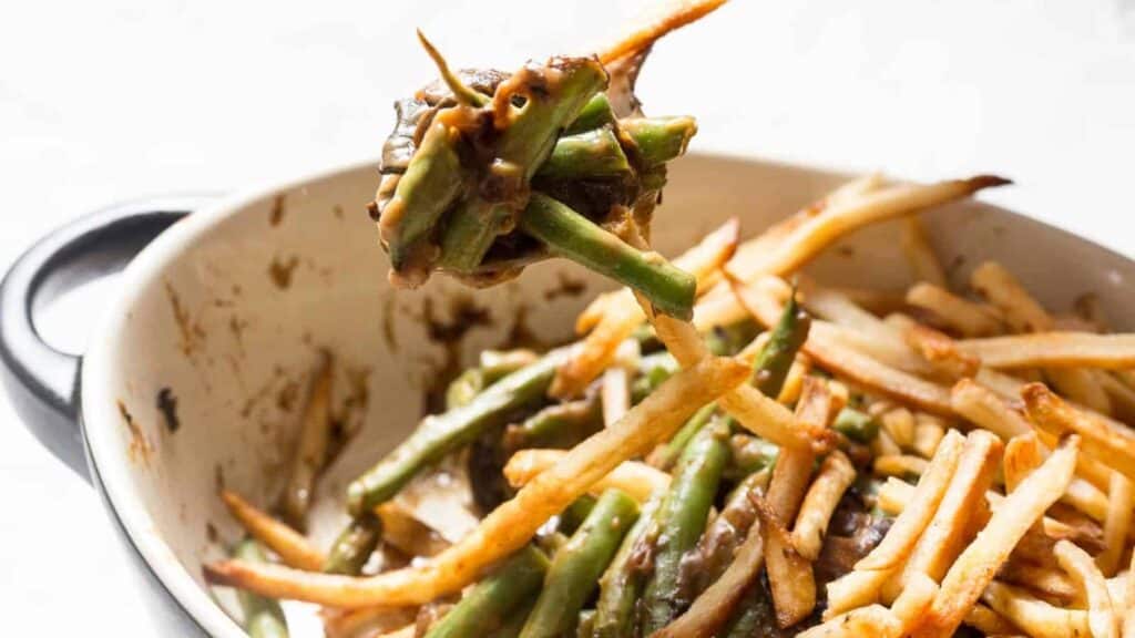 A casserole dish filled with cooked green beans and thin, crispy French fries, with a spoon lifting a portion of the mixture. Some fries and beans are coated in a dark sauce.