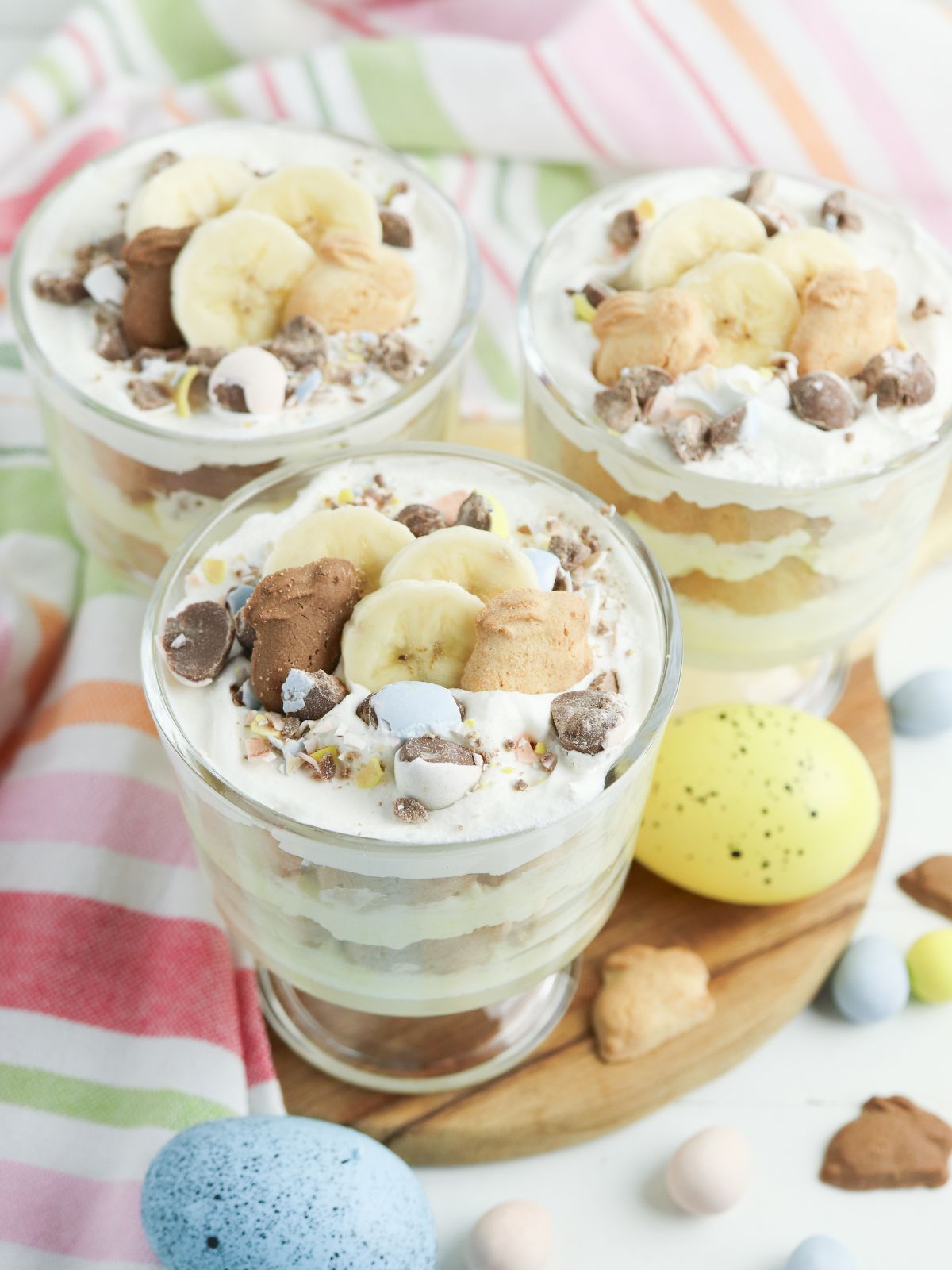 Three individual Easter banana pudding parfaits layered with vanilla pudding, bunny grahams, sliced bananas, whipped topping, and mini chocolate eggs on a wooden board.