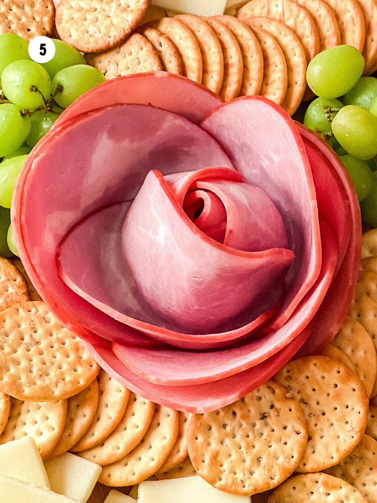 Ham rose arranged on a charcuterie board surrounded by crackers and grapes.