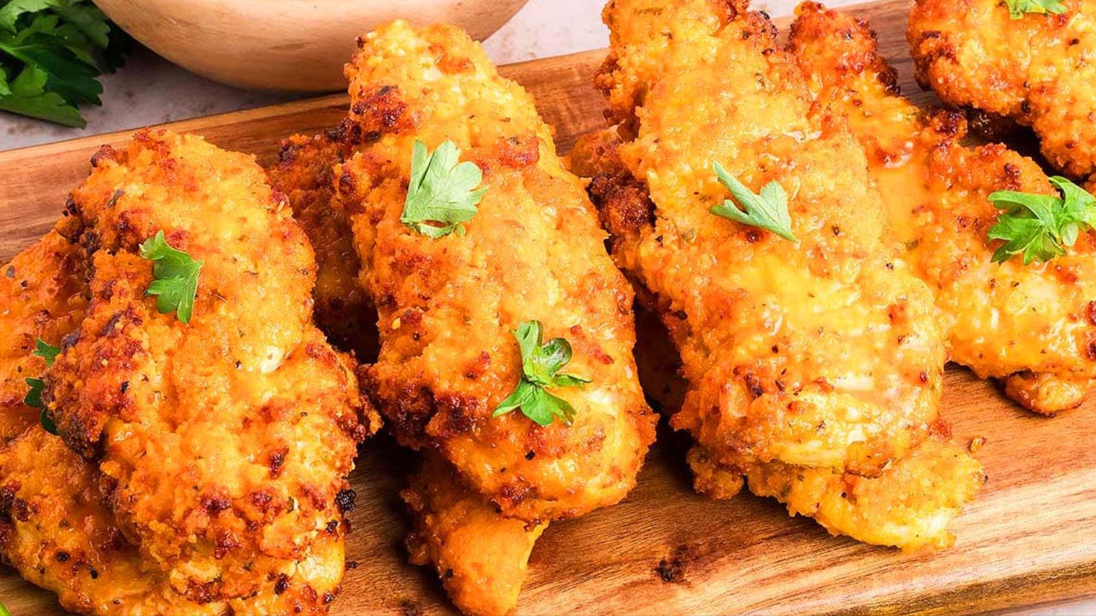 Cooked chicken tender pieces coated in a reddish sauce and arranged on a wooden board with small green herb leaves scattered on top.