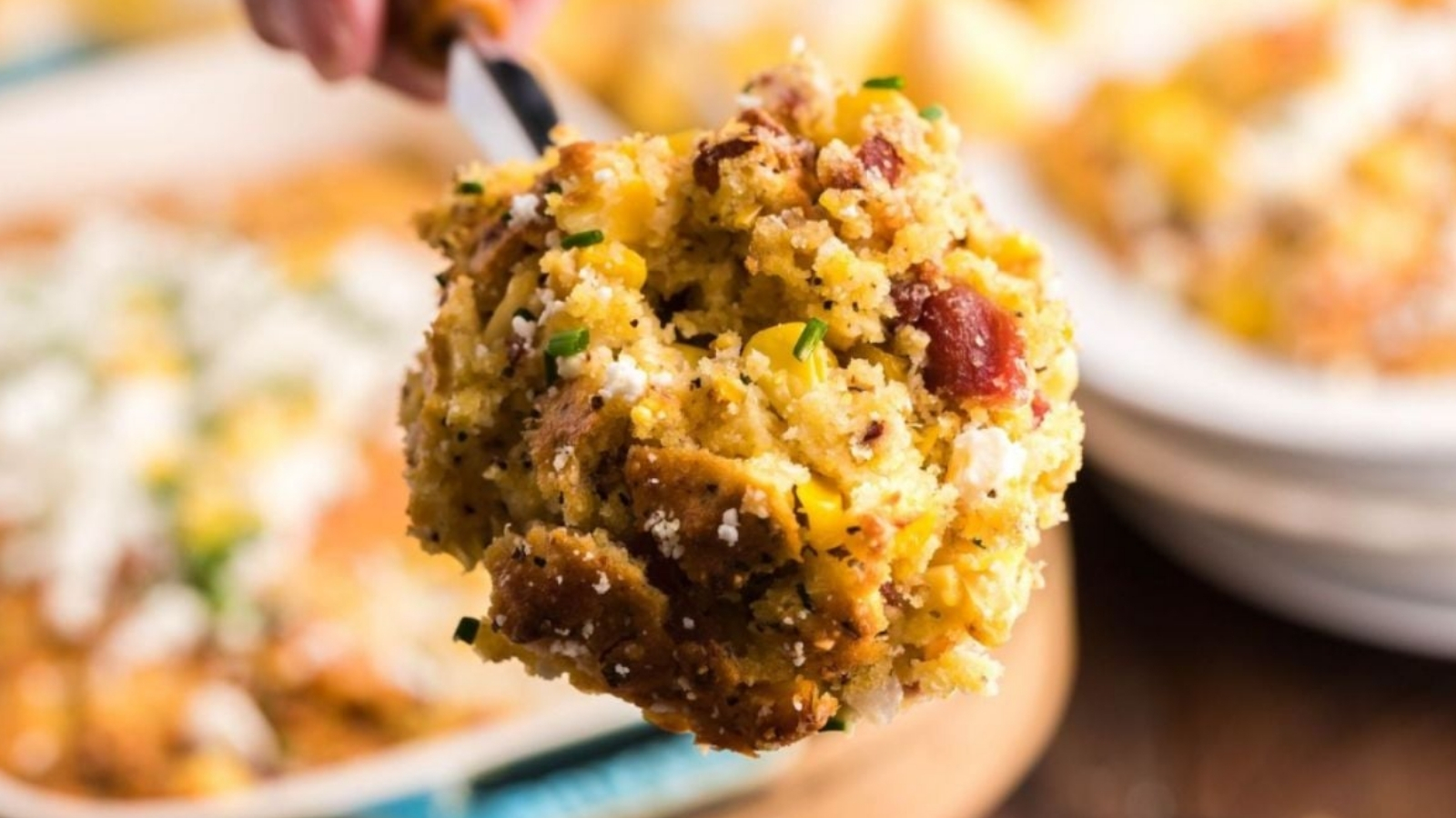 A spoon holds a scoop of cornbread stuffing with corn kernels, browned bread cubes, small meat pieces, chopped herbs, and grated cheese above a casserole dish.