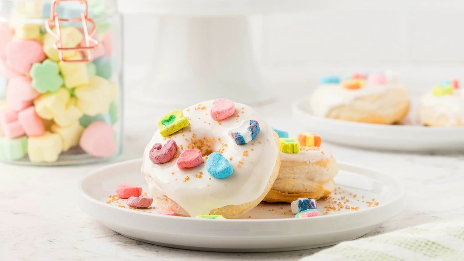 Donut with white icing topped with colorful cereal marshmallows and gold sprinkles on a white plate, with a jar of marshmallows in the background.