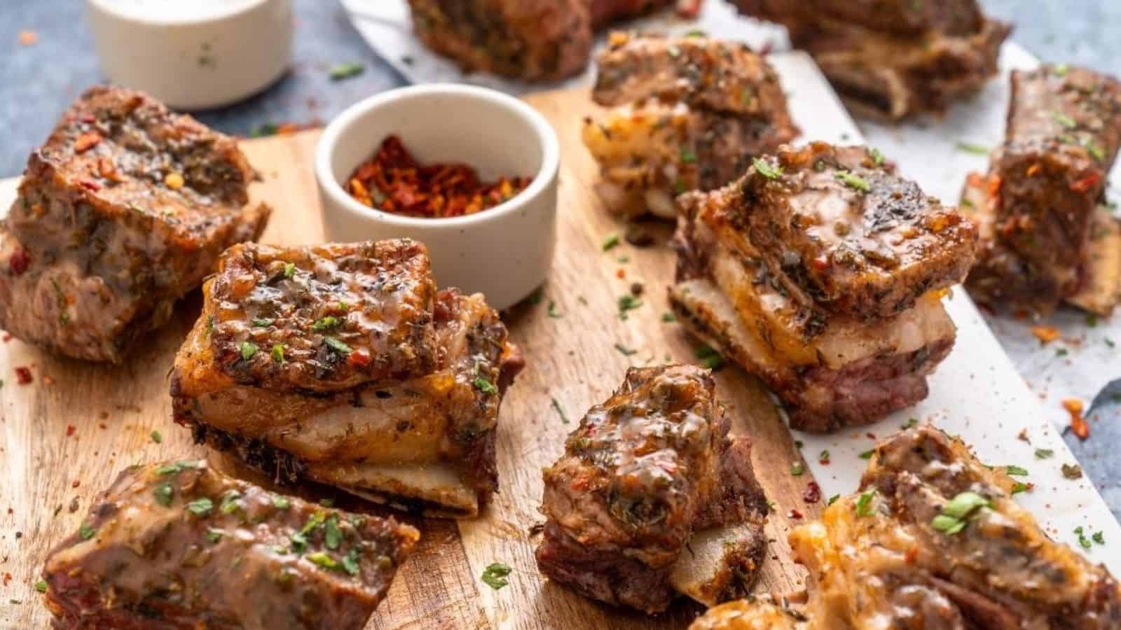 Several pieces of seasoned and cooked ribs on a wooden cutting board, garnished with herbs. Small bowls with spices are placed on the board.