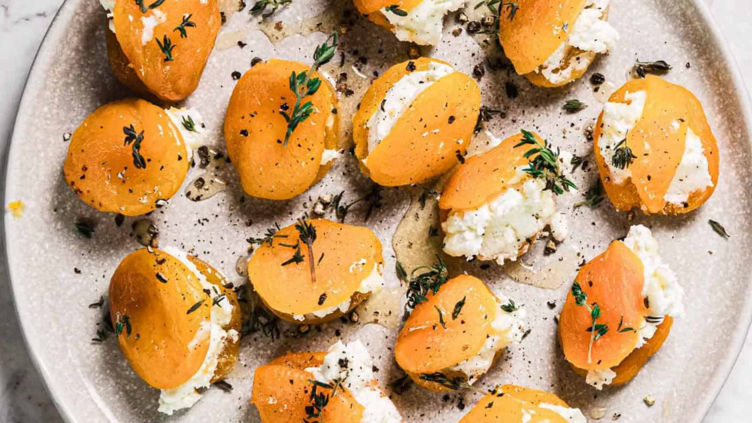 A plate of halved dried apricots stuffed with white cheese, garnished with fresh thyme and cracked black pepper, arranged on a round ceramic plate.
