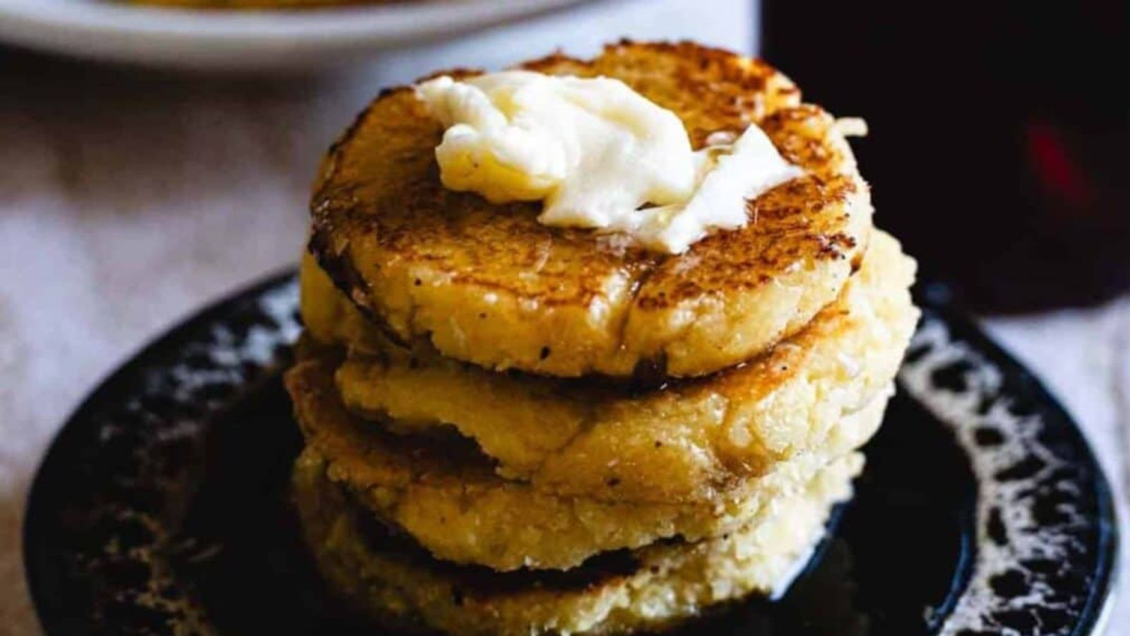 A stack of four golden brown cornmeal pancakes sits on a dark plate, topped with a dollop of melting butter.