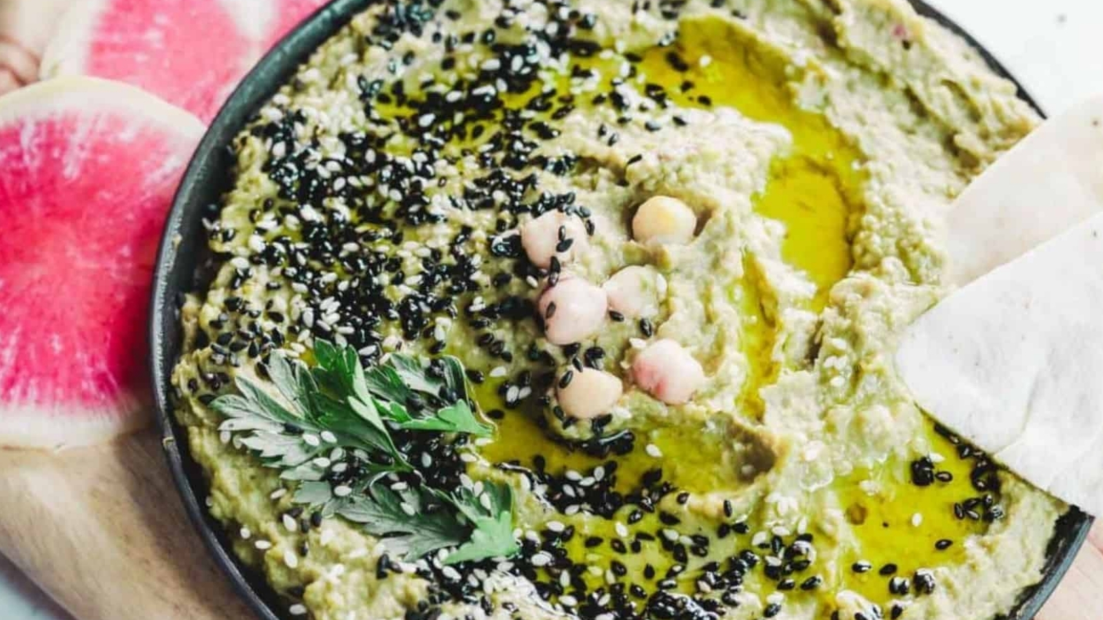 A bowl of hummus garnished with black sesame seeds, chickpeas, parsley, and olive oil, sits on a wooden board. Slices of pink and white watermelon radish are placed alongside.
