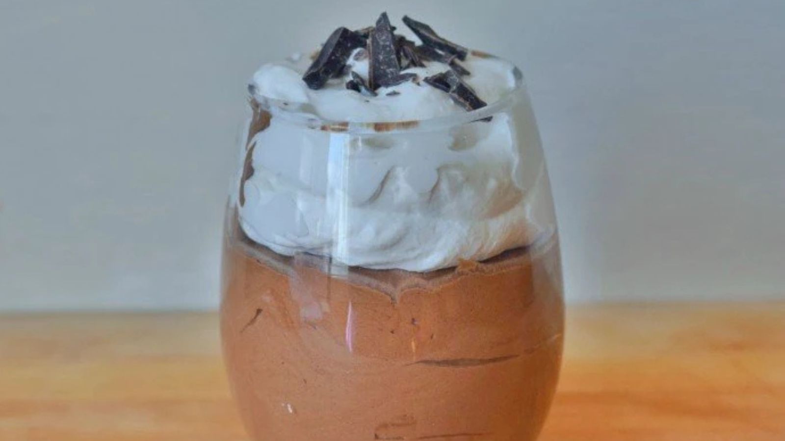 A glass filled with chocolate mousse topped with whipped cream and chocolate shavings, placed on a wooden surface.