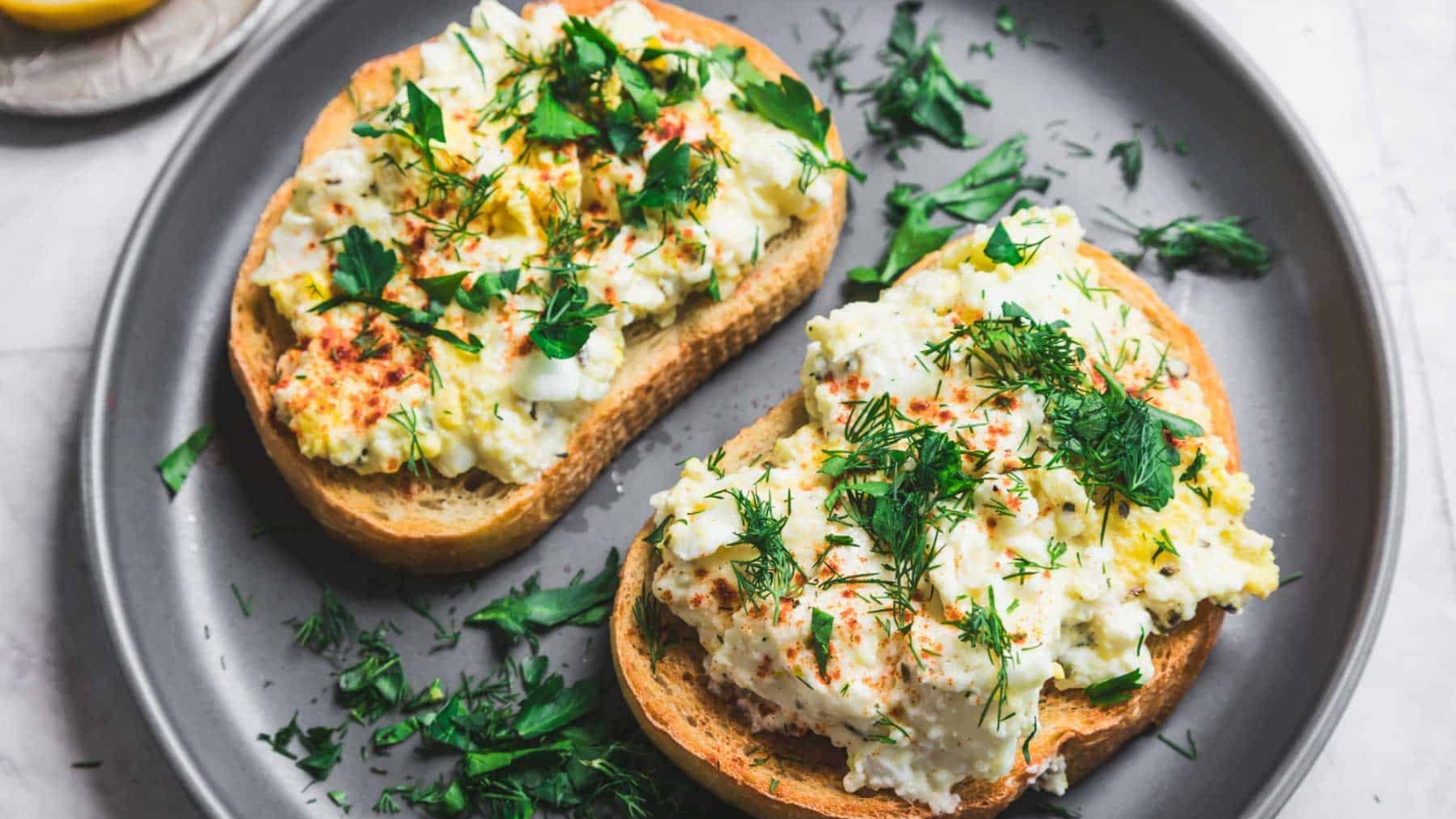 Two slices of bread topped with egg salad and garnished with fresh herbs are placed on a gray plate. A small dish with lemon slices is partially visible in the background on the left. The surface below is light-colored.