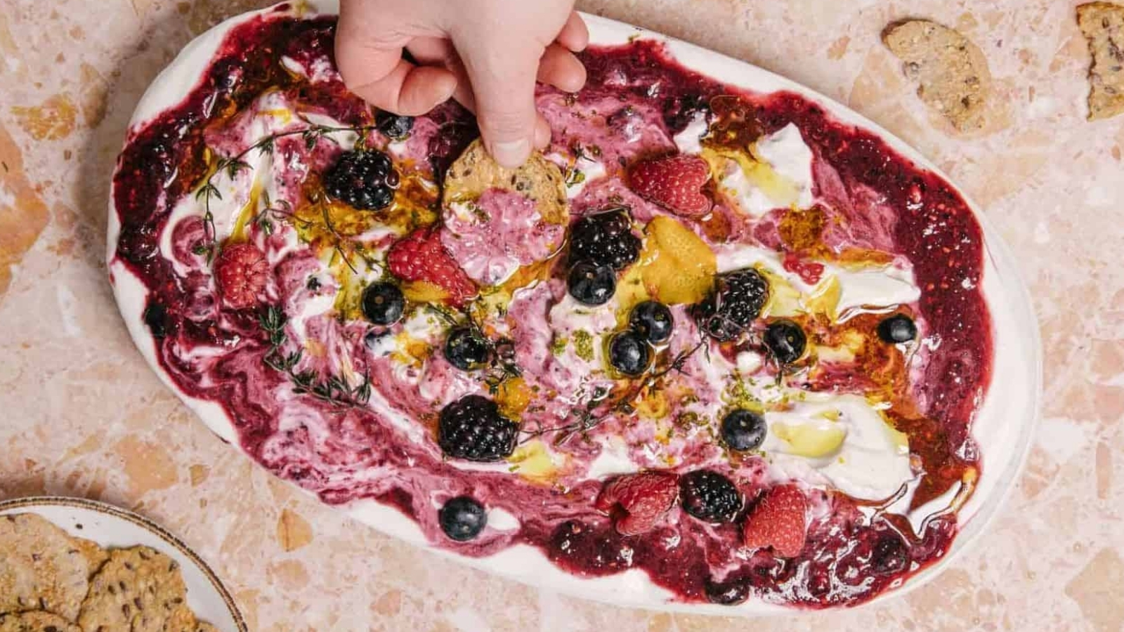 A hand dips a cracker into a platter of whipped cheese topped with a berry sauce, fresh berries, herbs, and olive oil, set on a beige marble surface. A plate with more crackers is visible in the corner.