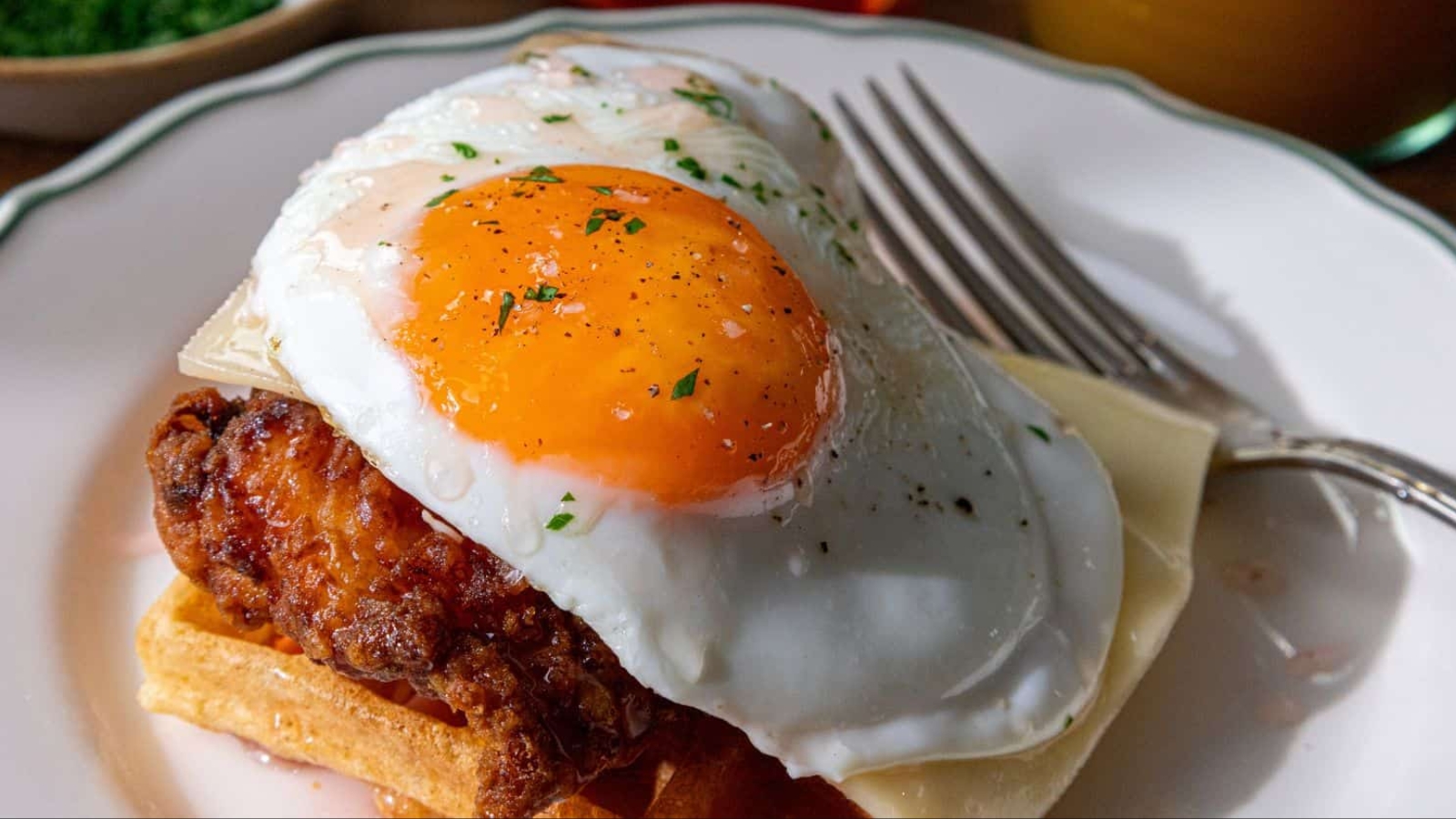 A close-up of a plate with a crispy waffle topped with fried chicken, melted cheese, and a sunny-side-up egg garnished with herbs. A fork rests on the plate's edge. In the background, there are blurred images of drinks and a small bowl.