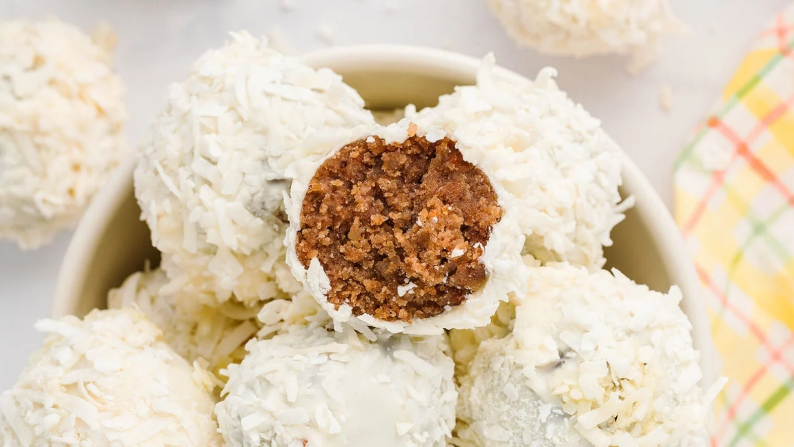 A bowl filled with coconut-coated balls, with one cut open to show a brown crumbly center, and additional balls placed around the bowl.