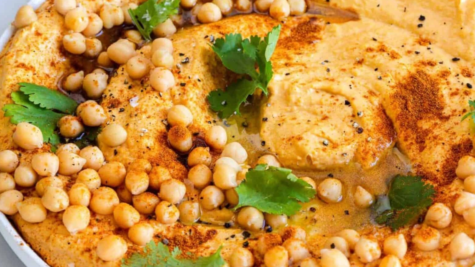A close-up of a bowl of hummus topped with chickpeas, olive oil, paprika, black pepper, and cilantro leaves. The hummus has a creamy texture and is visually garnished with spices and herbs.