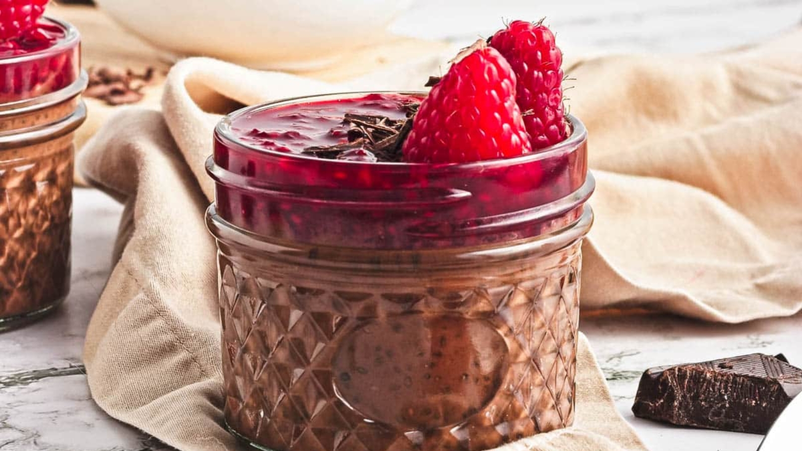 Two small jars filled with chocolate mousse, topped with raspberry sauce, fresh raspberries, and chocolate shavings. A beige cloth, chocolate pieces, loose raspberries, and a spoon are nearby on a marble surface.