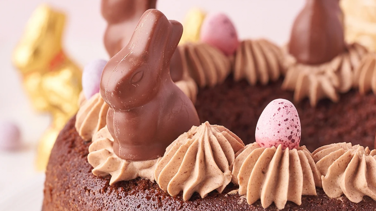 A chocolate bunny and speckled candy eggs sit on piped frosting dollops on top of a ring-shaped cake.