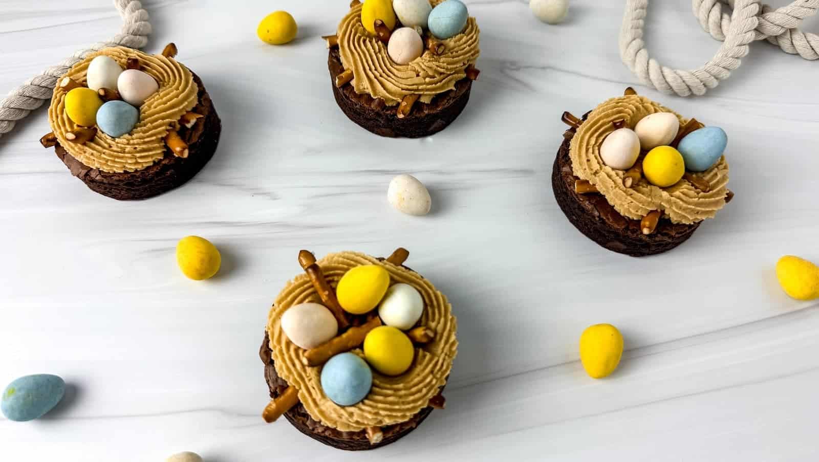 Four chocolate cupcakes topped with brown frosting, candy eggs, and pretzel sticks, arranged on a white surface with scattered colored candy eggs.