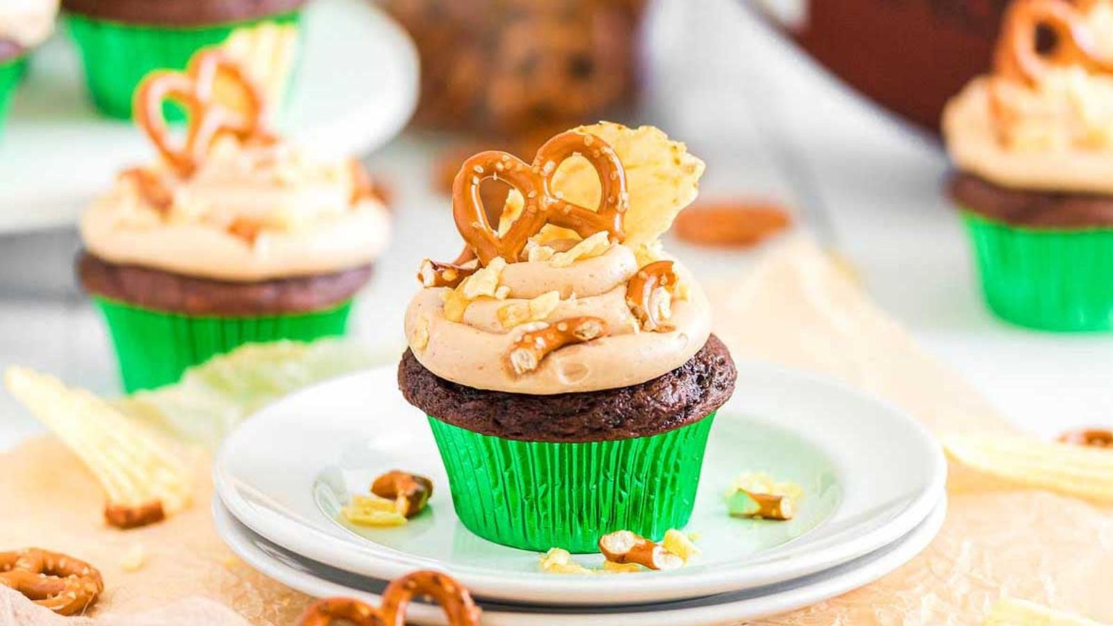 Chocolate cupcake with peanut butter frosting topped with pretzels and chopped peanuts in a green wrapper on a white plate.