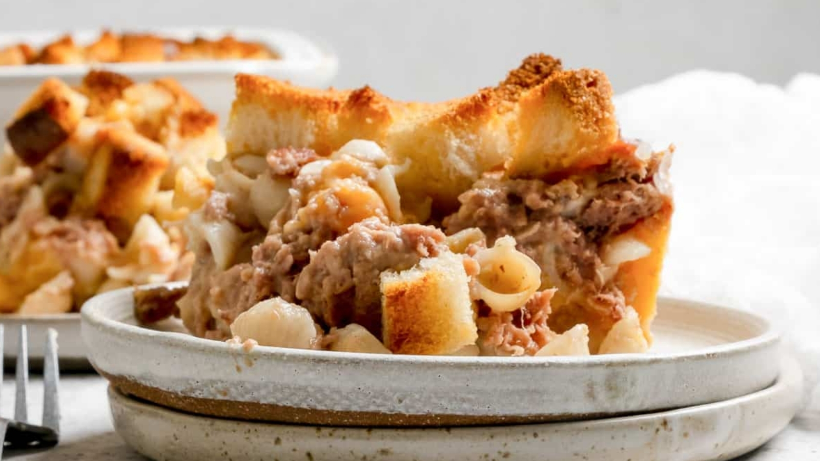A slice of baked casserole with pasta, meat, and toasted bread pieces served on a ceramic plate with another portion blurred in the background.