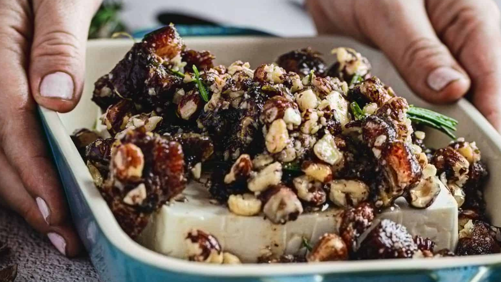 A block of soft white cheese topped with chopped nuts, dark sauce, and herbs in a rectangular dish held by hands.