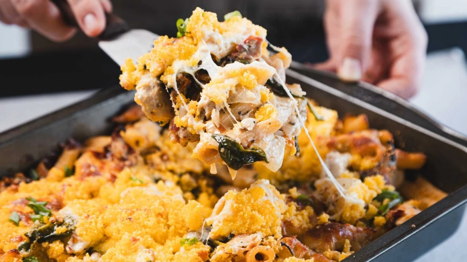 A hand lifts a portion of baked pasta with melted cheese from a casserole dish using a spatula.