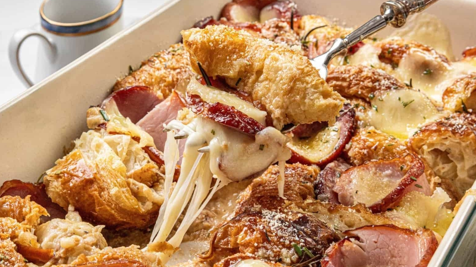 A fork lifts a portion of croissant breakfast casserole with cheese and slices of fruit, from a baking dish. A cup sits in the background.