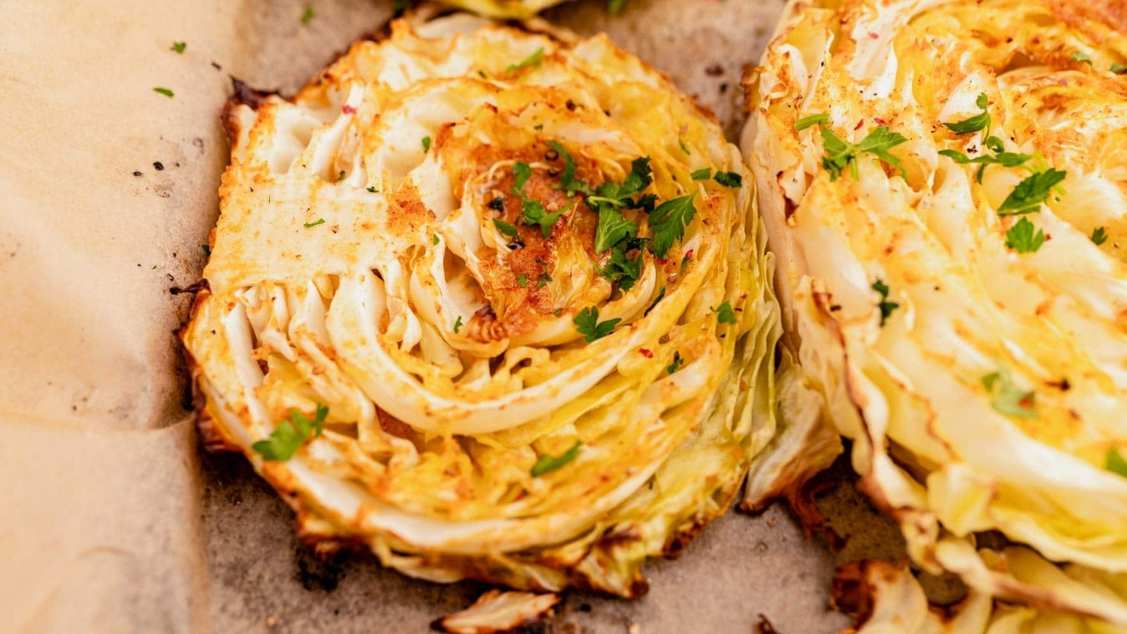 Two thick roasted cabbage slices with browned edges and chopped herbs on parchment paper.