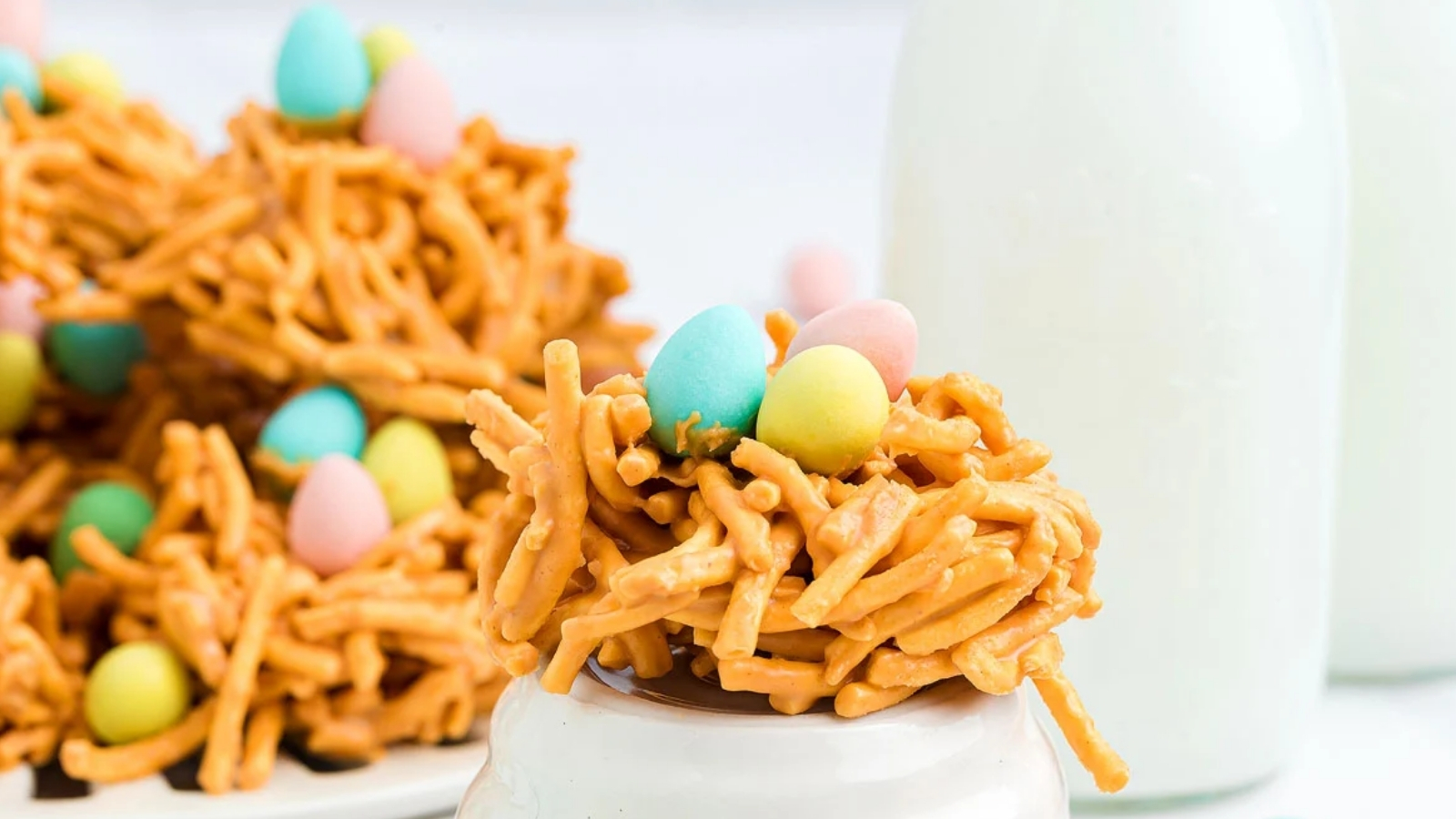 A pretzel snack cluster topped with pastel egg-shaped candies sits on an upside-down white cup with more clusters on a plate and a glass of milk in the background.