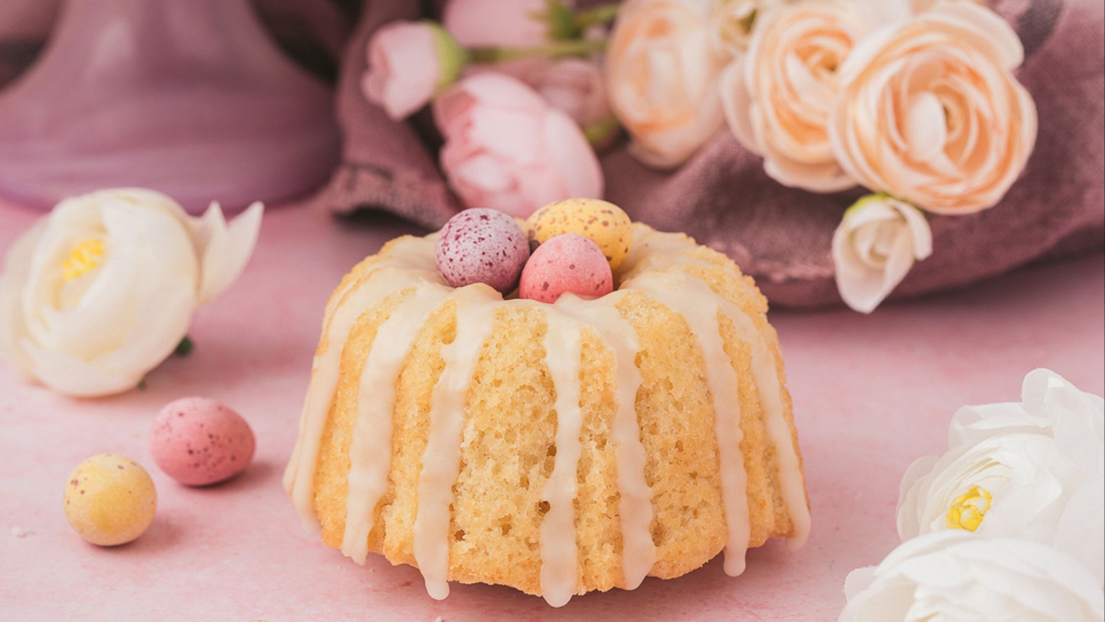 Mini Bundt cakes with white glaze topped with pastel chocolate eggs arranged on a surface.
