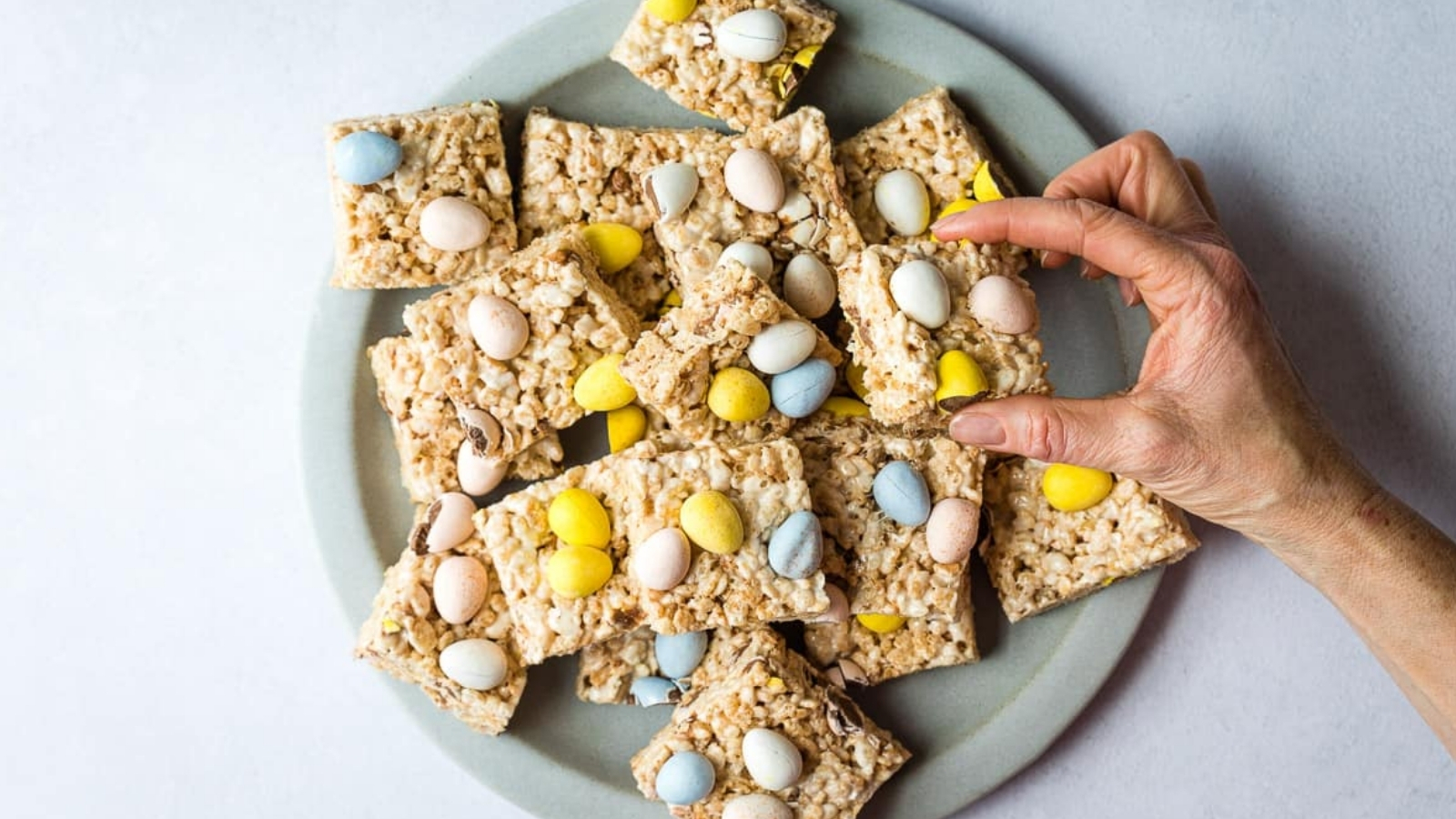 A hand holds a square cereal treat topped with small pastel candies above a plate filled with similar squares.