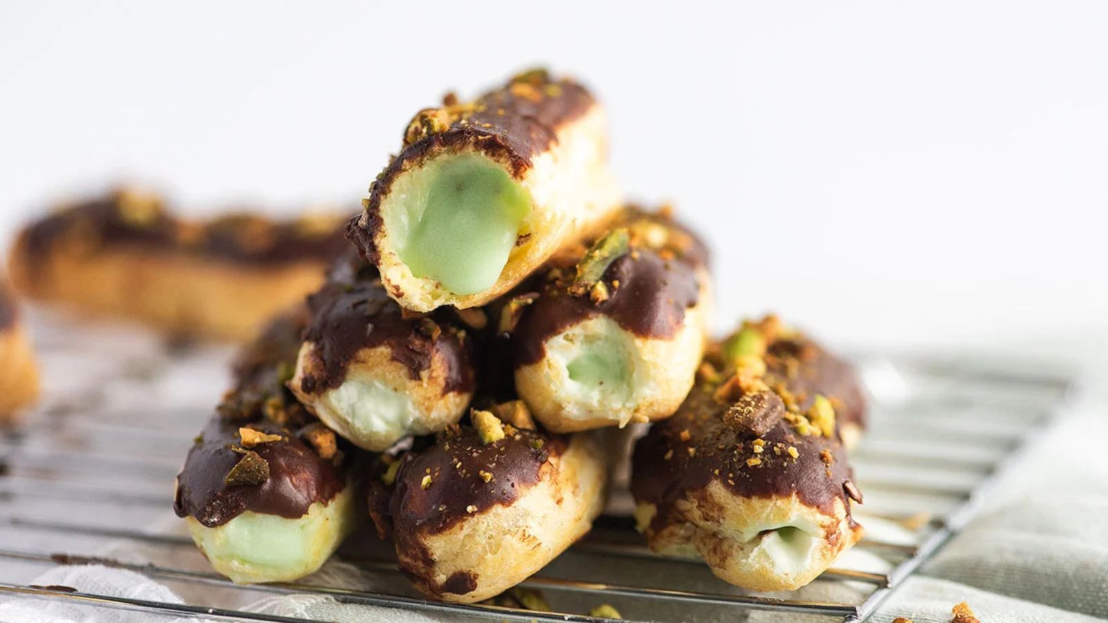 Stack of eclairs filled with green cream, topped with chocolate glaze and chopped nuts, resting on a metal cooling rack.