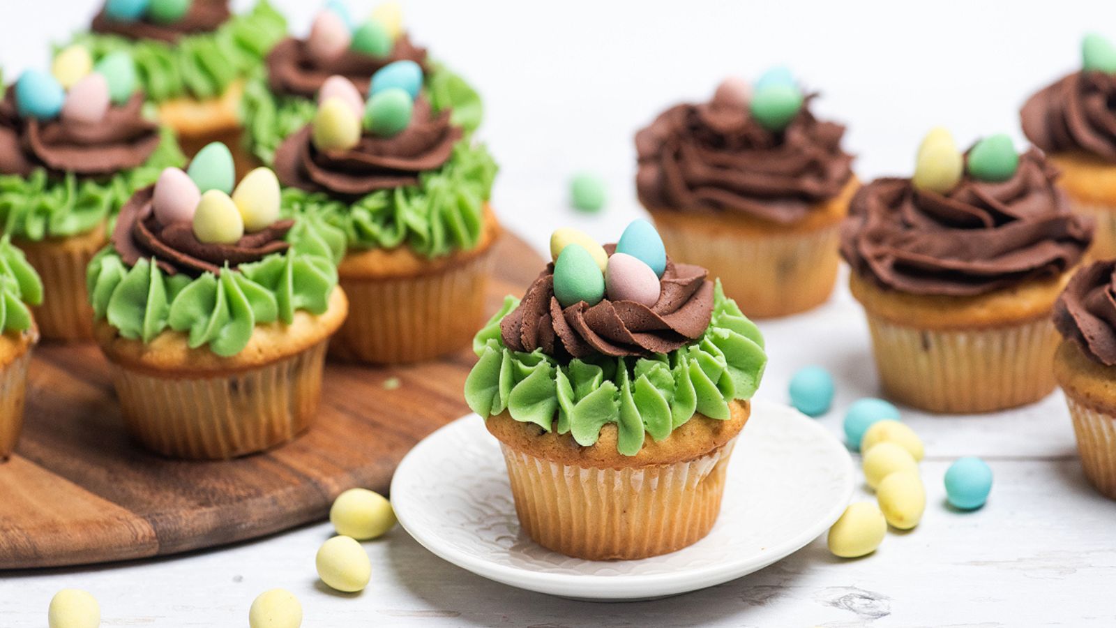 Cupcakes topped with green and brown frosting and small pastel candy eggs, with one cupcake on a white plate and others on a wooden board behind it.