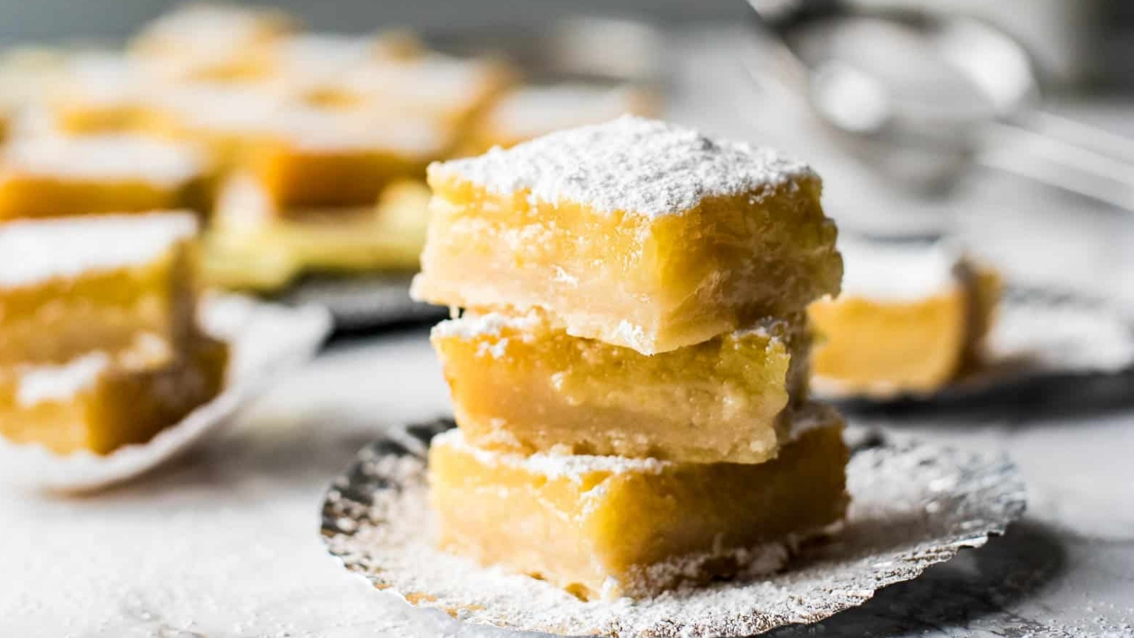 A stack of three lemon bars on a plate with more lemon bars in the background.