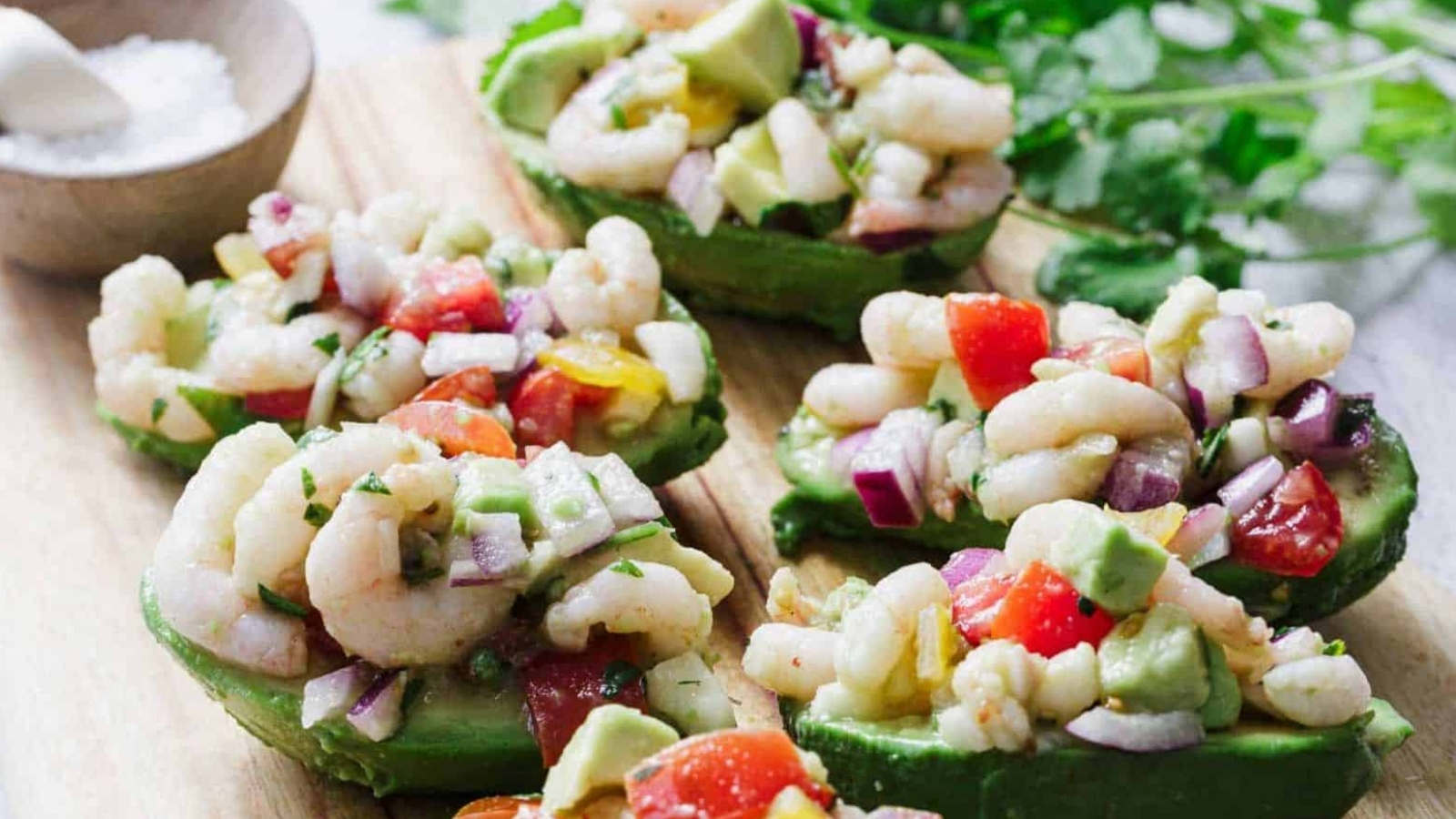 A black plate with a halved avocado filled with a shrimp salad, garnished with lime. A fork rests beside it. A wooden board holds more avocado halves in the background. A wooden bowl with mixed salad ingredients is partially visible.