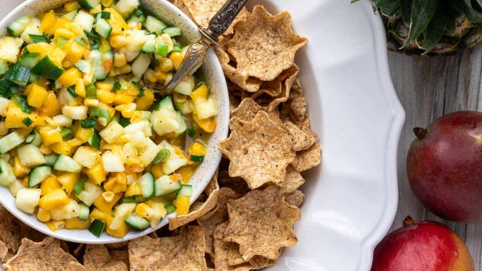 A bowl of chopped cucumber, corn, and yellow fruit with a spoon, surrounded by star-shaped tortilla chips on a large white plate, with two mangoes and a pineapple top nearby.