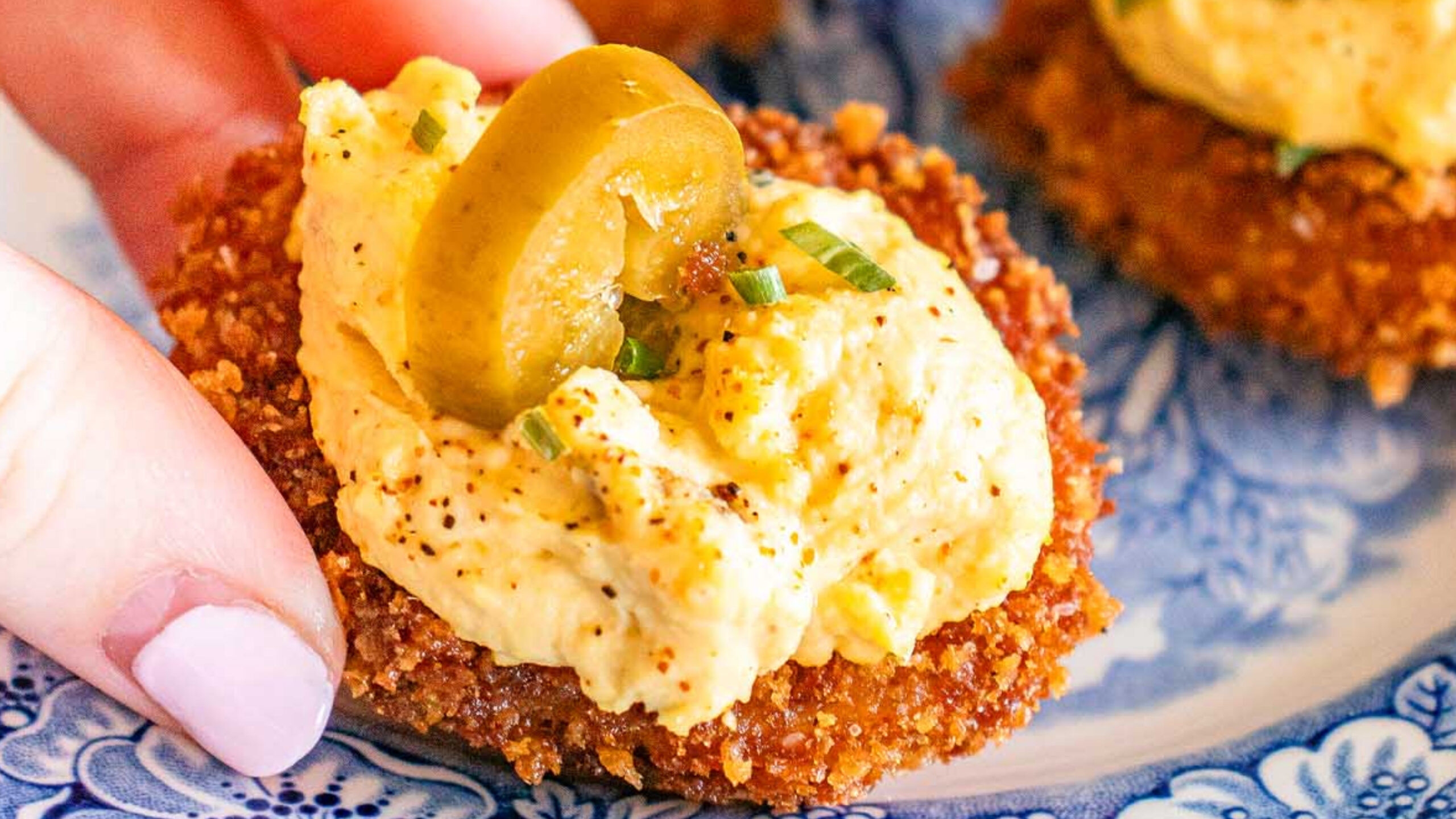 A hand holds a Fried Deviled Egg topped with a creamy spread and a sliced green olive on a blue patterned plate with two similar appetizers.
