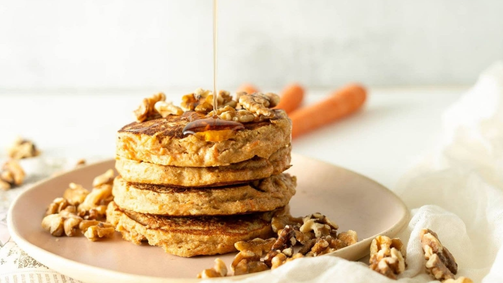 A stack of three pancakes topped with walnuts is on a pink plate. Syrup is being drizzled over the pancakes. Additional walnuts are scattered around the plate. Two carrots are partially visible in the background.