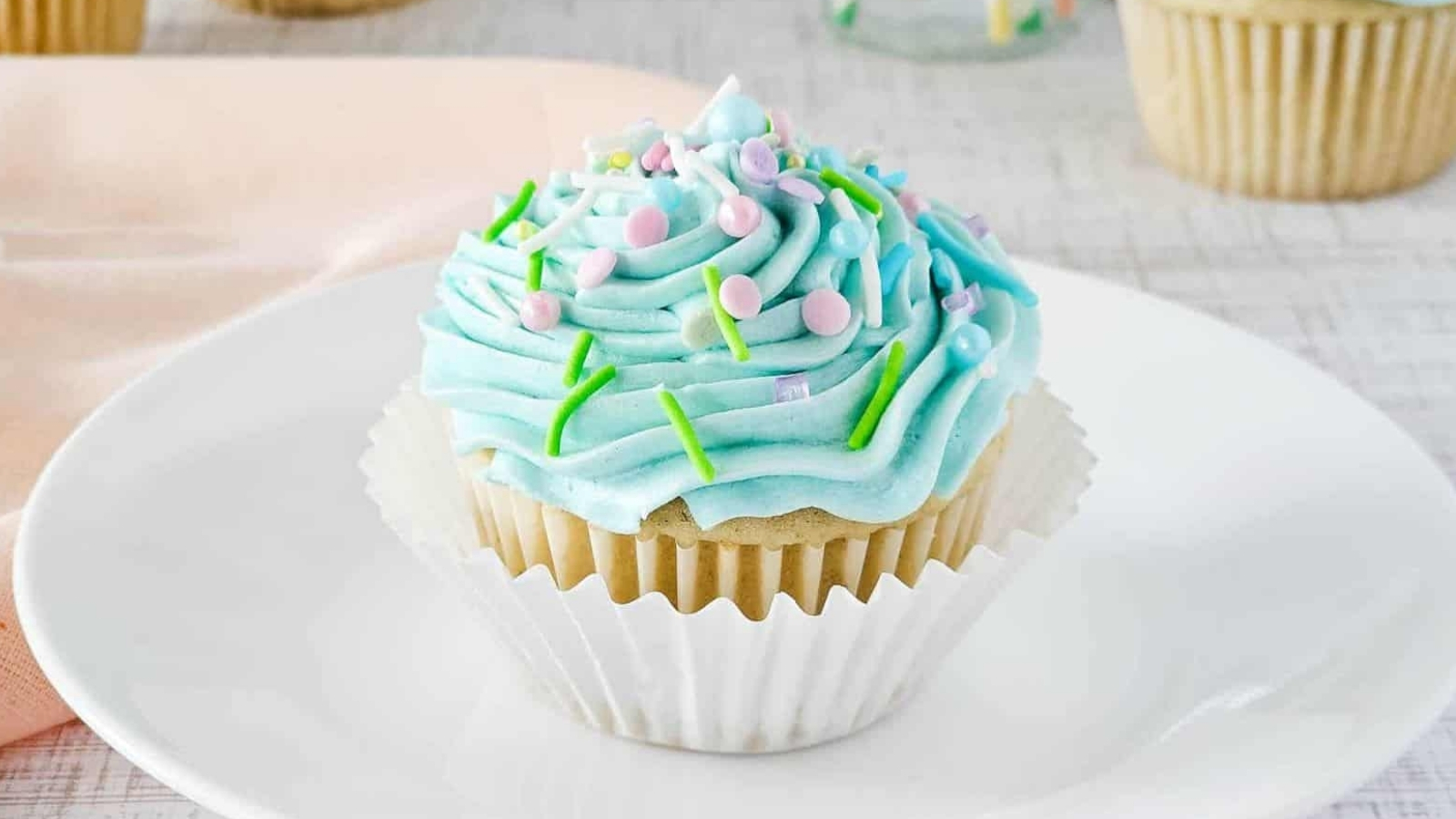 A vanilla cupcake with light blue frosting sits on a white plate. The frosting is topped with pastel sprinkles and green icing. The cupcake is in a white paper liner. A light pink cloth is partially visible in the background.