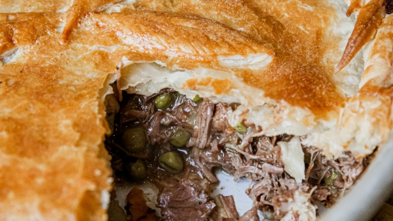 A white baking dish with a baked crust pie cut open to reveal shredded meat, peas, and carrot pieces, with a black fork resting on top.