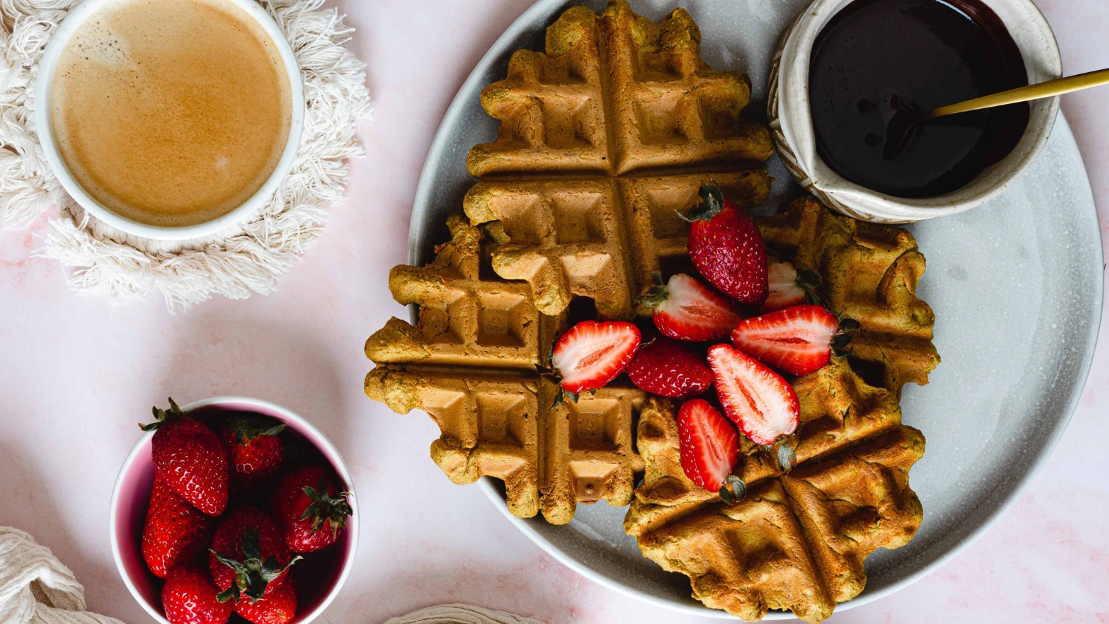 A plate of four golden brown waffles topped with halved strawberries. A small jug of syrup is placed next to the waffles. The plate is on a light-colored surface, with two folded cloth napkins nearby.