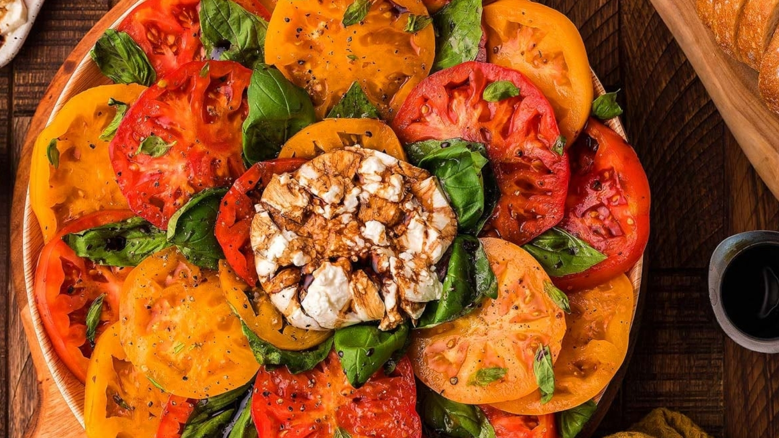 Sliced red and yellow tomatoes and basil leaves arranged around a ball of burrata drizzled with dark glaze on a round platter, with a small bowl of dark liquid and sliced bread at the side.