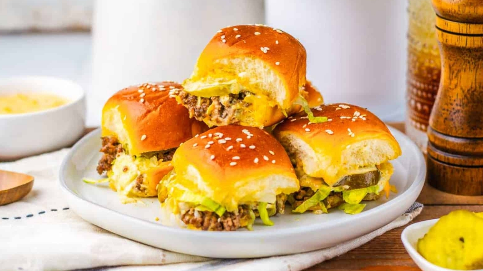 Four cheeseburger sliders with lettuce and pickles on a white plate, topped with sesame seed buns.