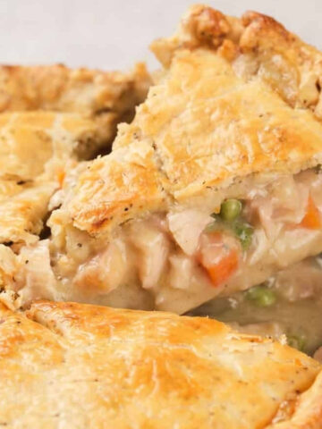 A serving of chicken pie with a golden crust on a white plate, showing pieces of chicken, peas, and carrots, with more pie in the background.