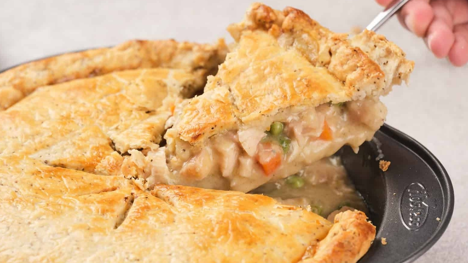 A serving of chicken pie with a golden crust on a white plate, showing pieces of chicken, peas, and carrots, with more pie in the background.