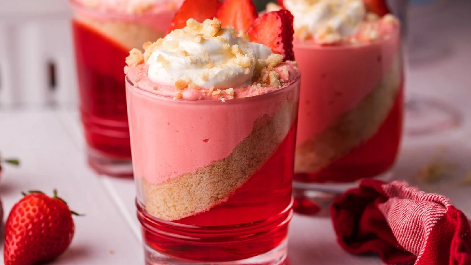 Three glasses filled with layered red gelatin, crumb base, and pink mousse, topped with whipped cream, crumbs, and sliced strawberries.