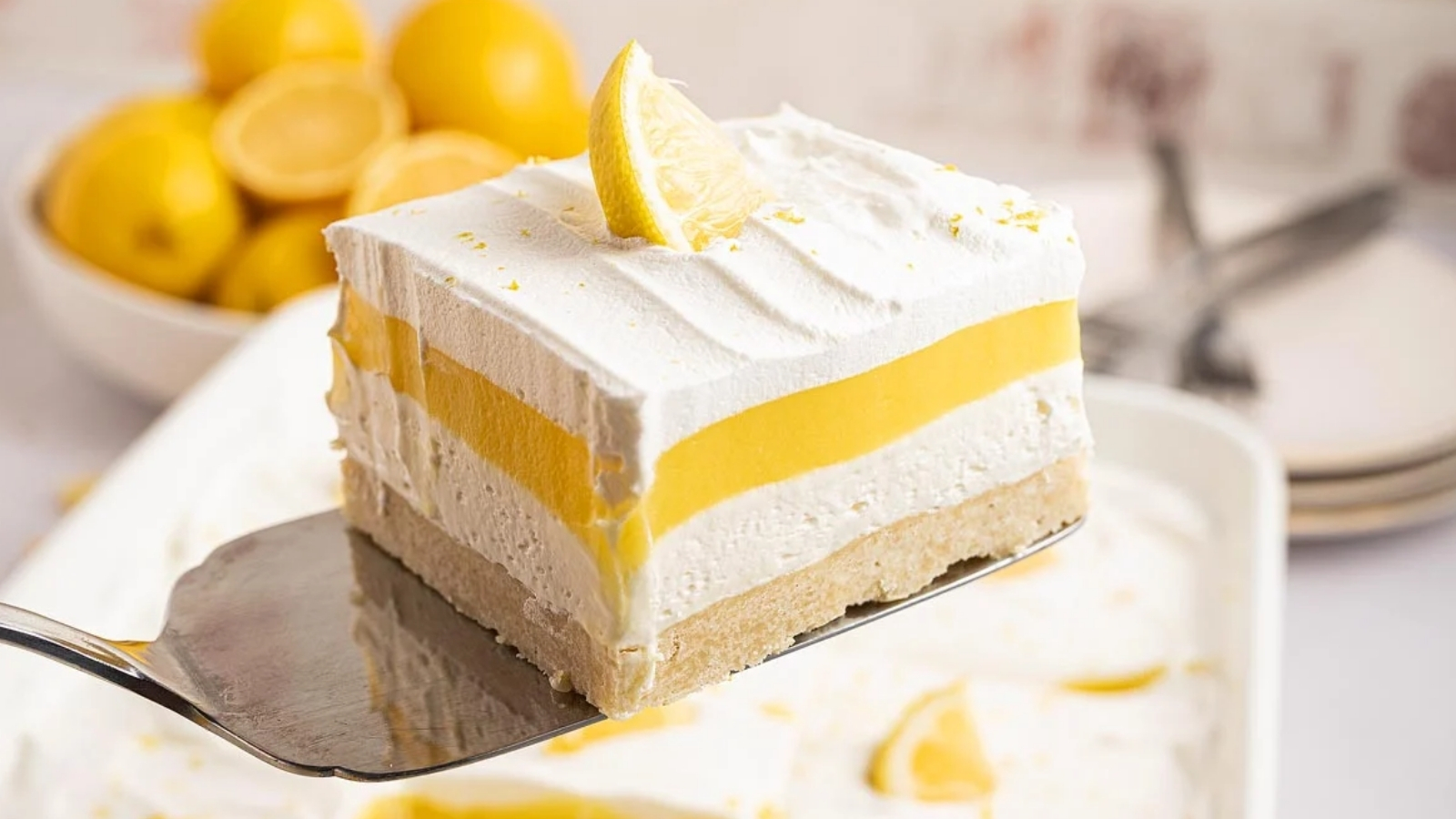 A square slice of layered lemon dessert with a crust, creamy filling, lemon layer, and whipped topping sits on a spatula, with whole lemons visible in the background.